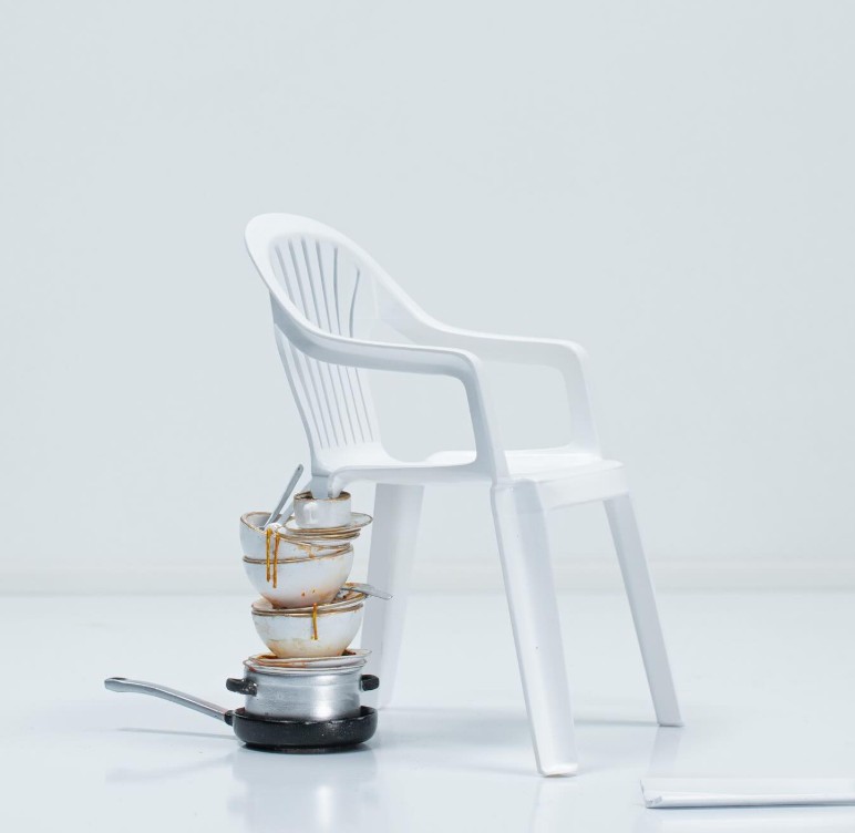 A miniature white monobloc armchair with one broken leg, propped up on a stack of dirty dishes, cups and pans, the snapped leg lying on the floor beside it